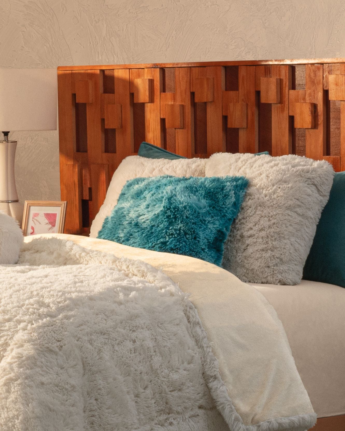 A cozy bed with a wooden geometric headboard, white and beige bedding, and Intima Hogar’s Juego De Cojín Everest in gray and teal microfibra. A small framed picture sits on the bedside table next to a modern lamp.