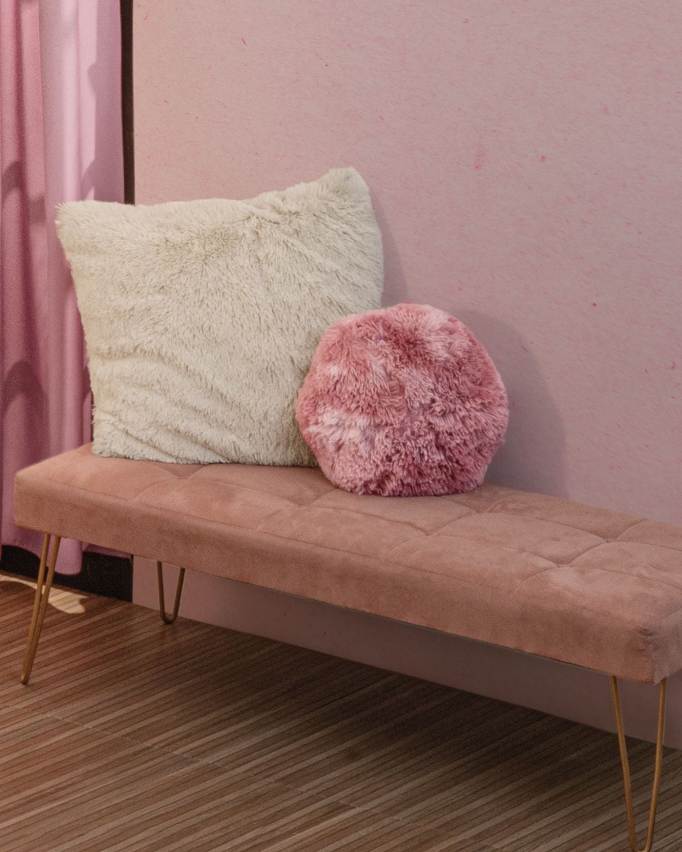 A pink velvet bench with gold legs sits against a pink wall. On the bench are two plush pillows: one large and white, the other a Cojín de Lujo Flamenco Ball from Intima Hogar. The floor is brown with a striped pattern.