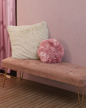 A pink velvet bench with gold legs sits against a pink wall. On the bench are two plush pillows: one large and white, the other a Cojín de Lujo Flamenco Ball from Intima Hogar. The floor is brown with a striped pattern.