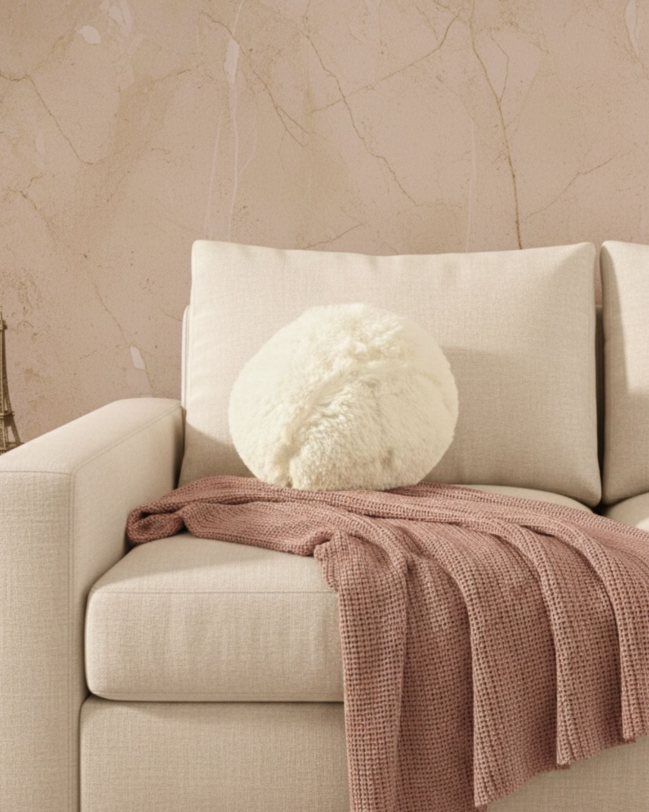 A beige armchair with an Intima Hogar Cojín de Lujo Iglú Ball in white and a textured pink throw blanket on the armrest, set against a light brown marble-patterned wall.