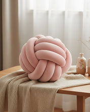 A soft Cojín Lazo Rosa by Intima Hogar rests on a beige knitted throw atop a wooden table. Two decorative vases and sheer white curtains in the background highlight this original accent piece.