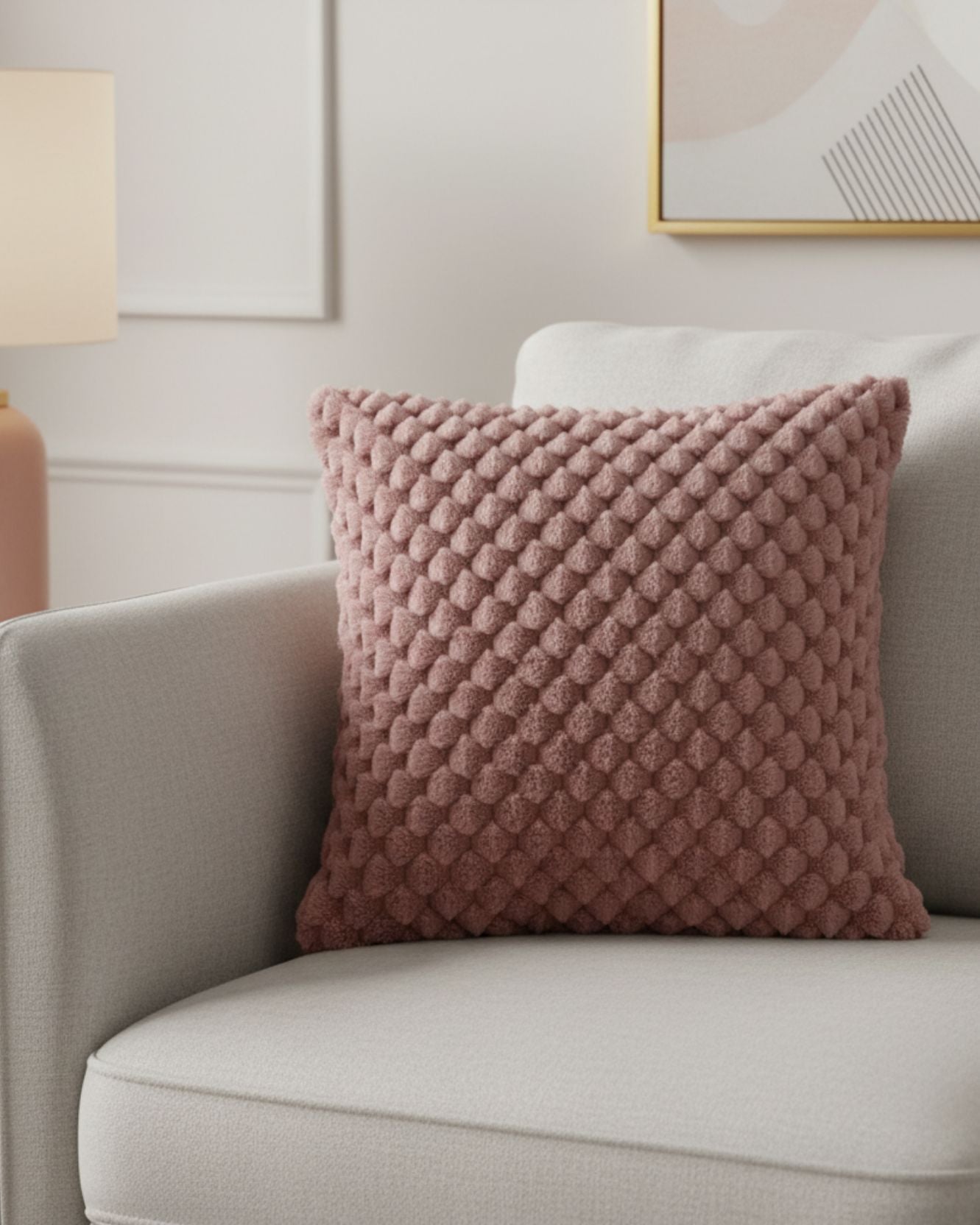 El Cojín de Lujo Aruba de Intima Hogar, en tono malva y con textura de puntos en relieve, decora un sillón gris claro en una sala moderna con luz suave.