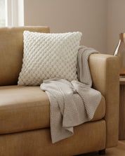 A beige sofa with a smooth texture, featuring the Cojín Nickel by Intima Hogar in white and a knitted beige throw, is beside a small wooden side table with a gold vase in a softly lit room.