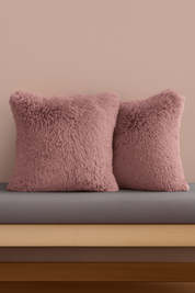 Two pink, fluffy Cojines de Lujo Liverpool by Intima Hogar are placed side by side on a gray cushion against a light pink wall, resting on a wooden bench.