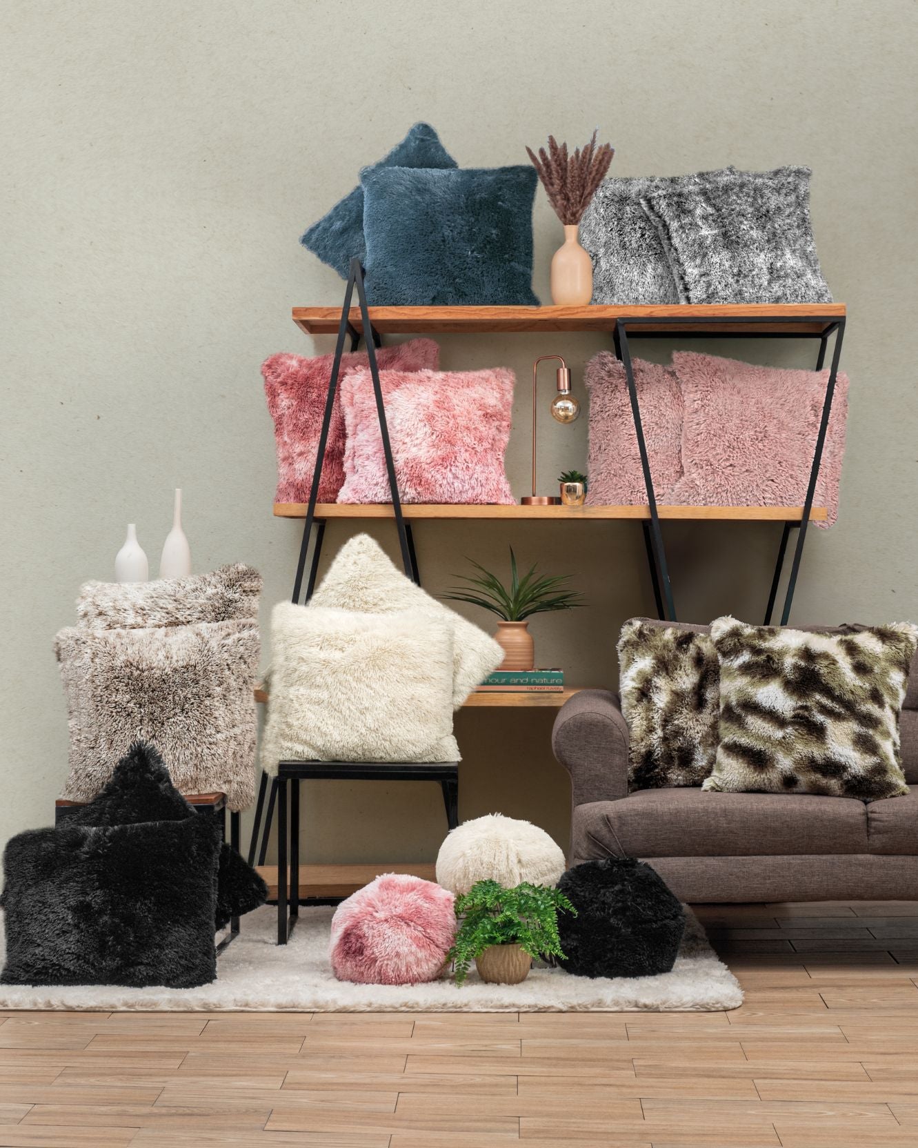 A cozy living room with shelves and a couch featuring Intima Hogar's Cojín de Lujo Albania Ball in pink, cream, black, blue, and gray with textura esponjosa—accented by decorative vases, books, and lush indoor plants.