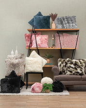 A cozy living room with shelves and a couch featuring Intima Hogar's Cojín de Lujo Albania Ball in pink, cream, black, blue, and gray with textura esponjosa—accented by decorative vases, books, and lush indoor plants.