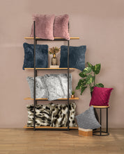 A black metal and wood shelving unit displays Cojines de Lujo Liverpool by Intima Hogar in pink, blue, gray, and animal print. Nearby, a small round table holds red and gray pillows beside a green potted plant on a wooden floor.