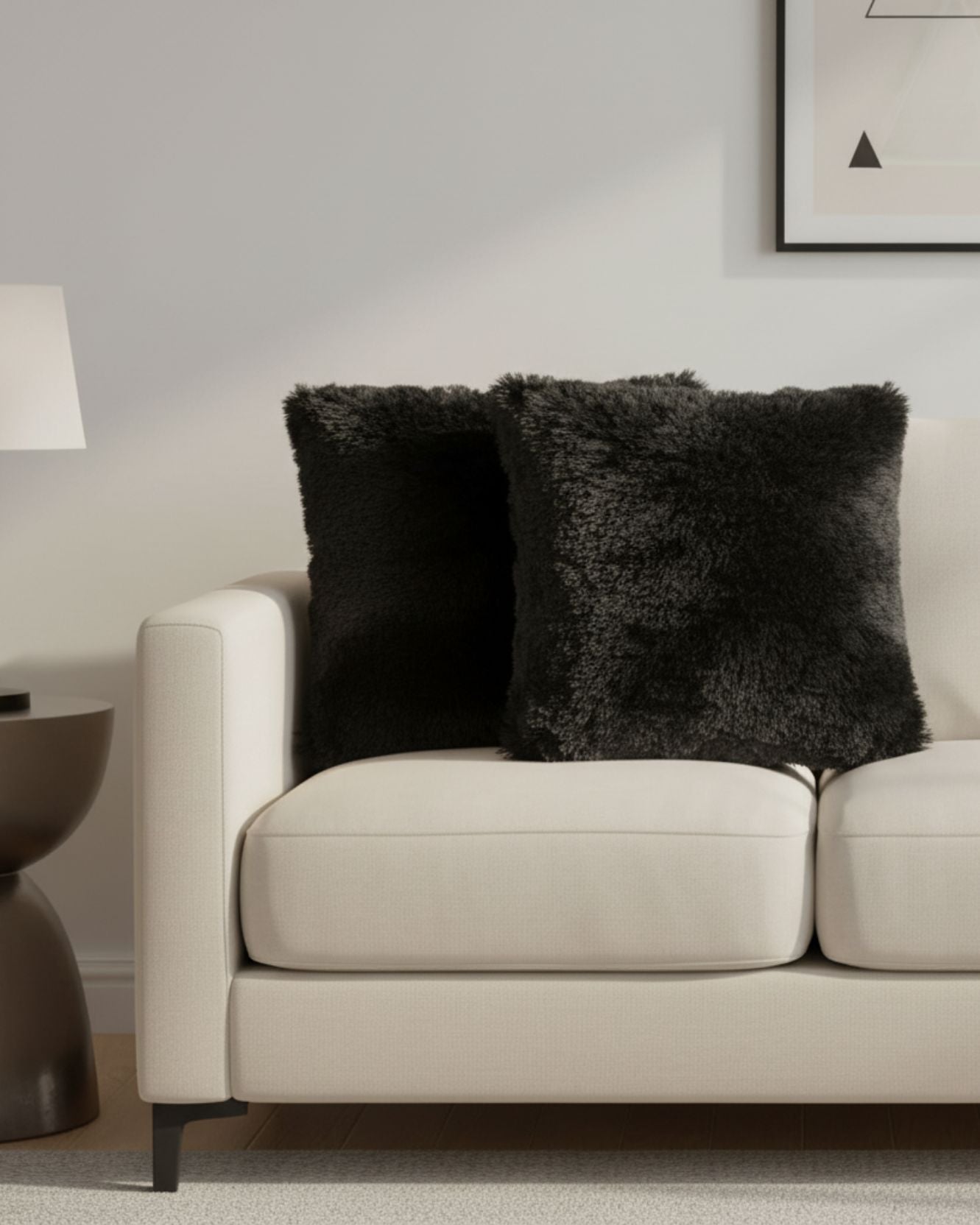 A modern living room with a cream-colored sofa accented by the Juego de Cojines Albania by Intima Hogar, a round dark side table with a white lamp, and part of framed wall art above the sofa.