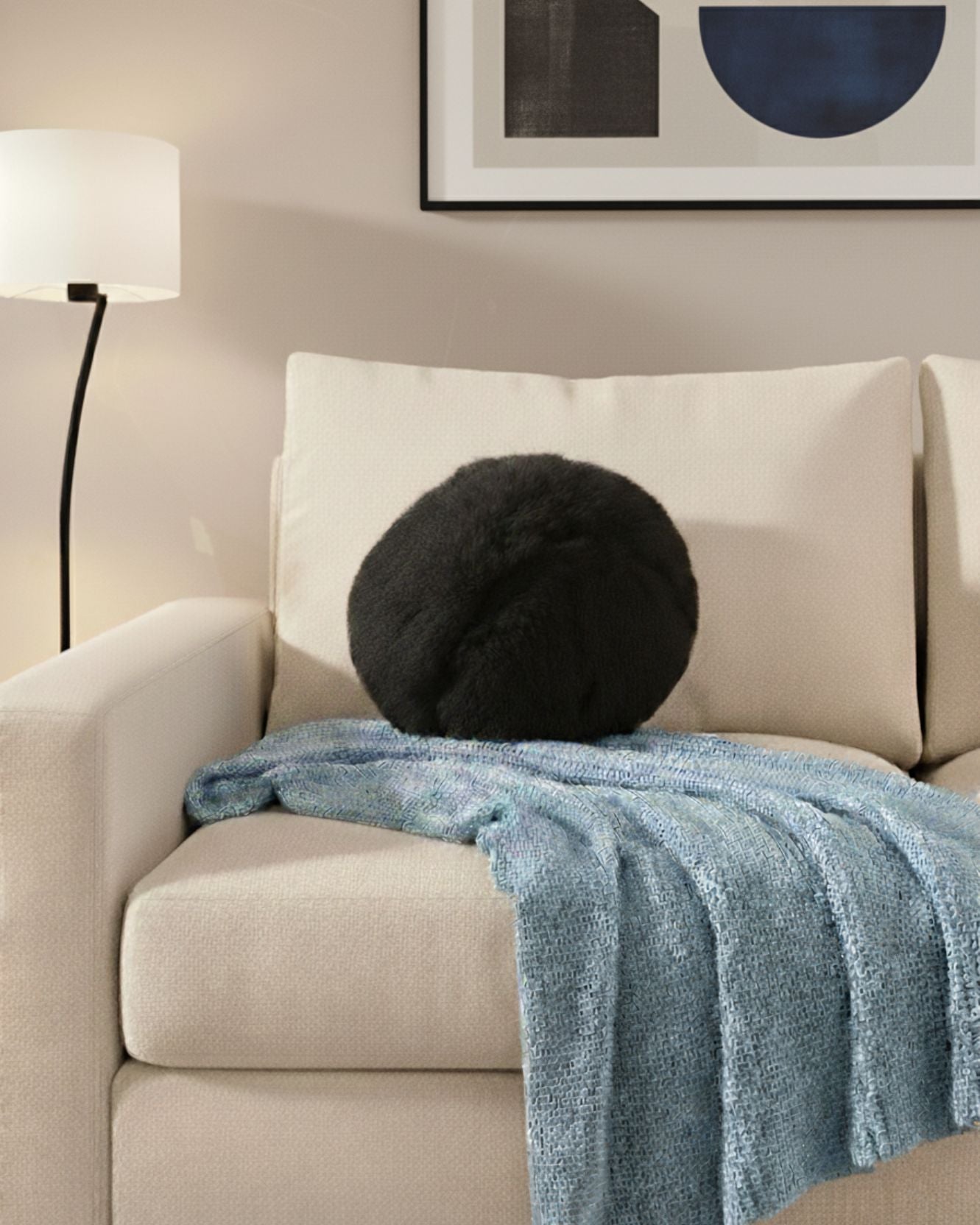 A beige armchair with the Intima Hogar Cojín de Lujo Albania Ball black round pillow and a light blue knitted throw sits by a floor lamp beneath framed abstract art on a light-colored wall.