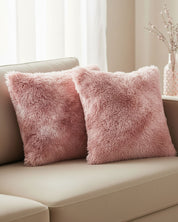 Dos cojines del Juego de Cojines de Lujo Flamenco de Intima Hogar, en tono rosa y textura peludita, están sobre un sofá beige. De fondo, un florero con ramas claras decora una mesa cerca de una ventana con cortinas blancas translúcidas.