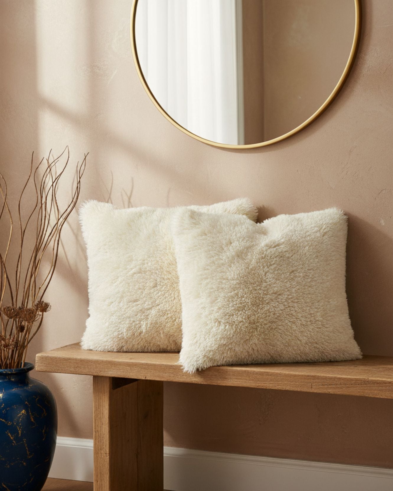 Dos cojines decorativos Juego de Cojines de Lujo Iglú de Intima Hogar, color crema y textura suave, están sobre una banca de madera bajo un espejo redondo dorado. Un florero azul con ramas secas añade un acabado luminoso sobre la pared beige.