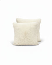 Dos cojines cuadrados off-white del Juego de Cojines de Lujo Iglú de Intima Hogar, con textura suave y esponjosa, están apilados y se superponen ligeramente sobre un fondo blanco.