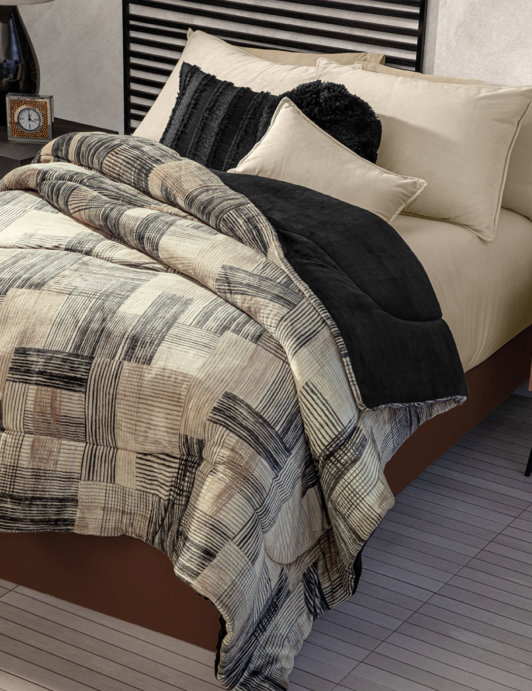 A neatly made bed with Intima Hogar's Coordinado de Cobertor Digital Soft Big Ben, beige sheets, black and cream decorative pillows. The room includes a striped headboard and a bedside table with a clock.