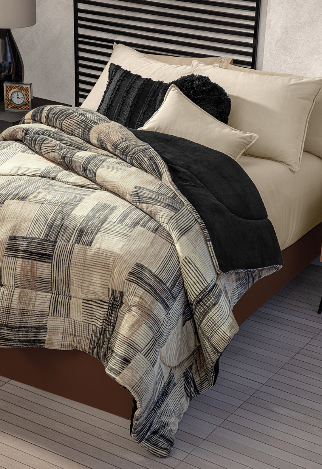 A neatly made bed with Intima Hogar's Coordinado de Cobertor Digital Soft Big Ben, beige sheets, black and cream decorative pillows. The room includes a striped headboard and a bedside table with a clock.