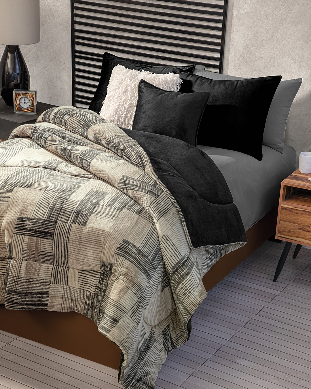 A neatly made bed features gray sheets, the Funda de Almohada Ensueño Negro by Intima Hogar on black pillows, a fluffy white cushion, a patterned beige and gray quilt, a modern black headboard, and a bedside clock for an inviting habitación.