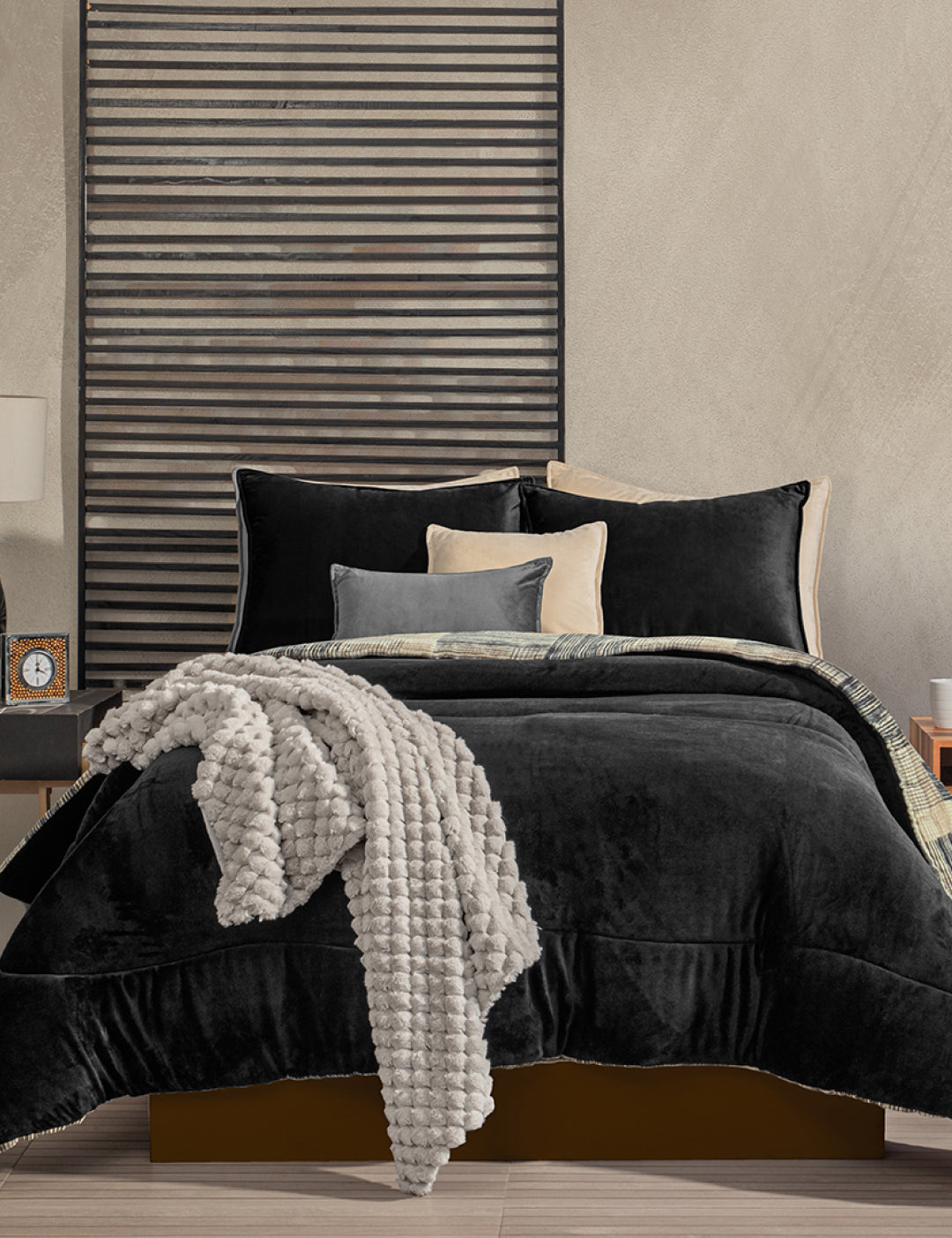 A modern bedroom features a neatly made bed with the Intima Hogar Coordinado de Cobertor Digital Soft Big Ben, black sheets, cream and gray pillows, a black slatted wall behind the headboard, and a nightstand with a clock and books.