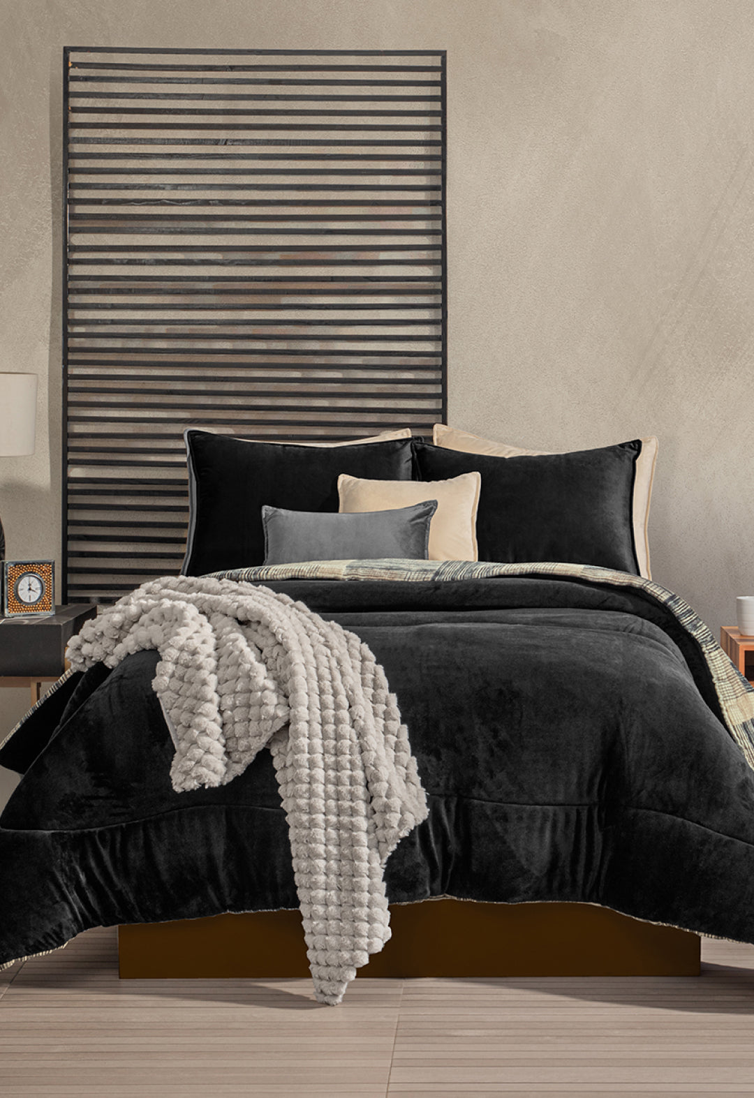 A modern bedroom features a neatly made bed with the Intima Hogar Coordinado de Cobertor Digital Soft Big Ben, black sheets, cream and gray pillows, a black slatted wall behind the headboard, and a nightstand with a clock and books.