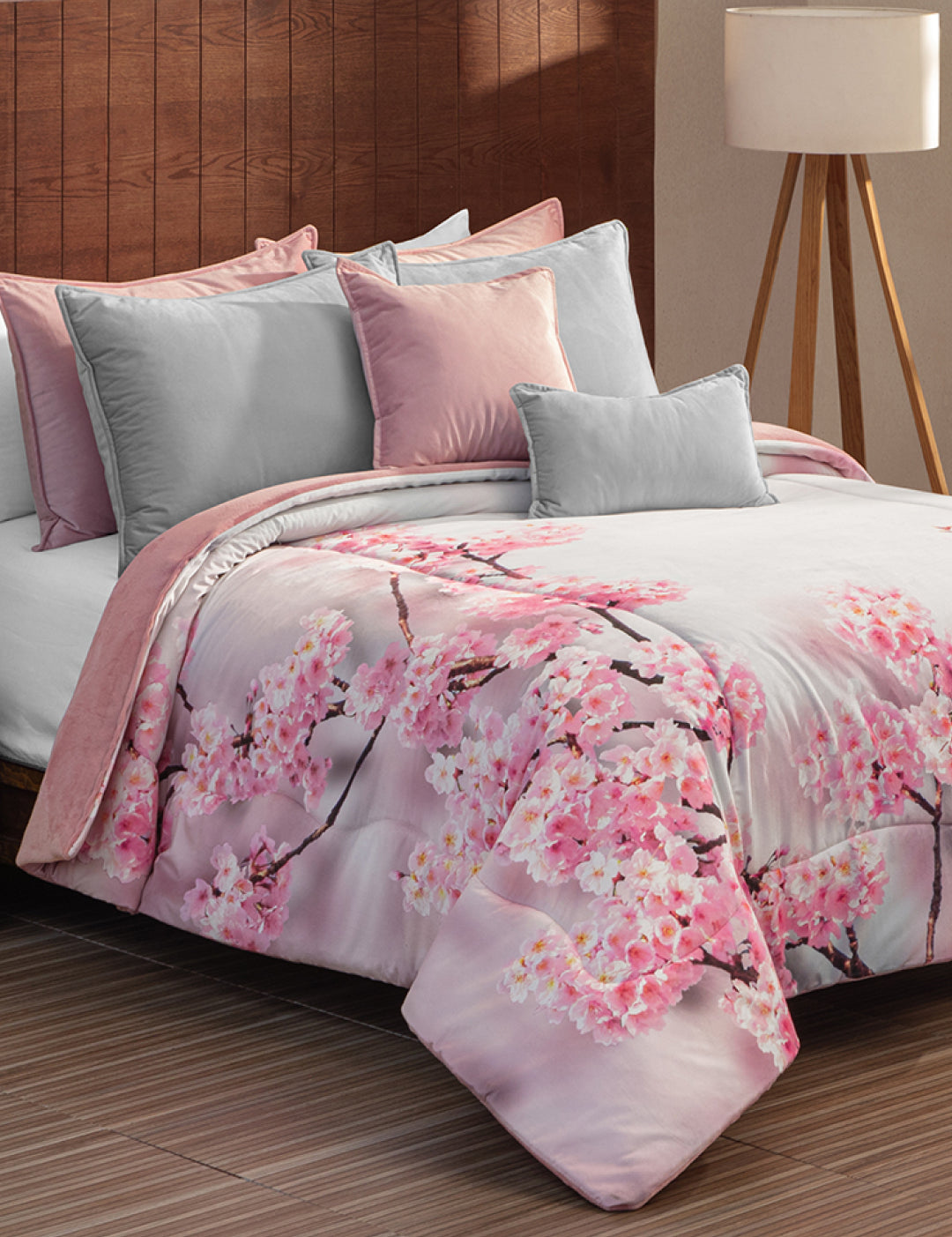 A bed features the Coordinado de Cobertor Digital Soft Florecer by Intima Hogar, with a pink and gray cherry blossom pattern, matching pillows, and a wooden headboard. A white-shade floor lamp stands in the background.