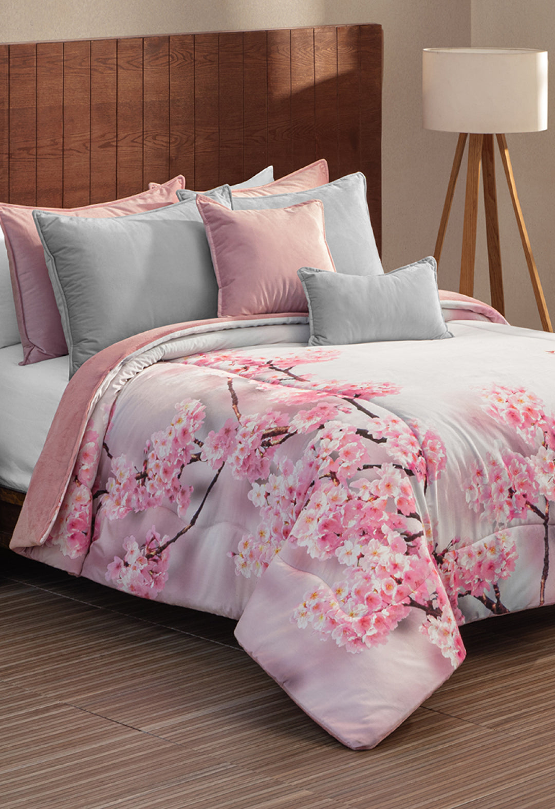 A bed features the Coordinado de Cobertor Digital Soft Florecer by Intima Hogar, with a pink and gray cherry blossom pattern, matching pillows, and a wooden headboard. A white-shade floor lamp stands in the background.