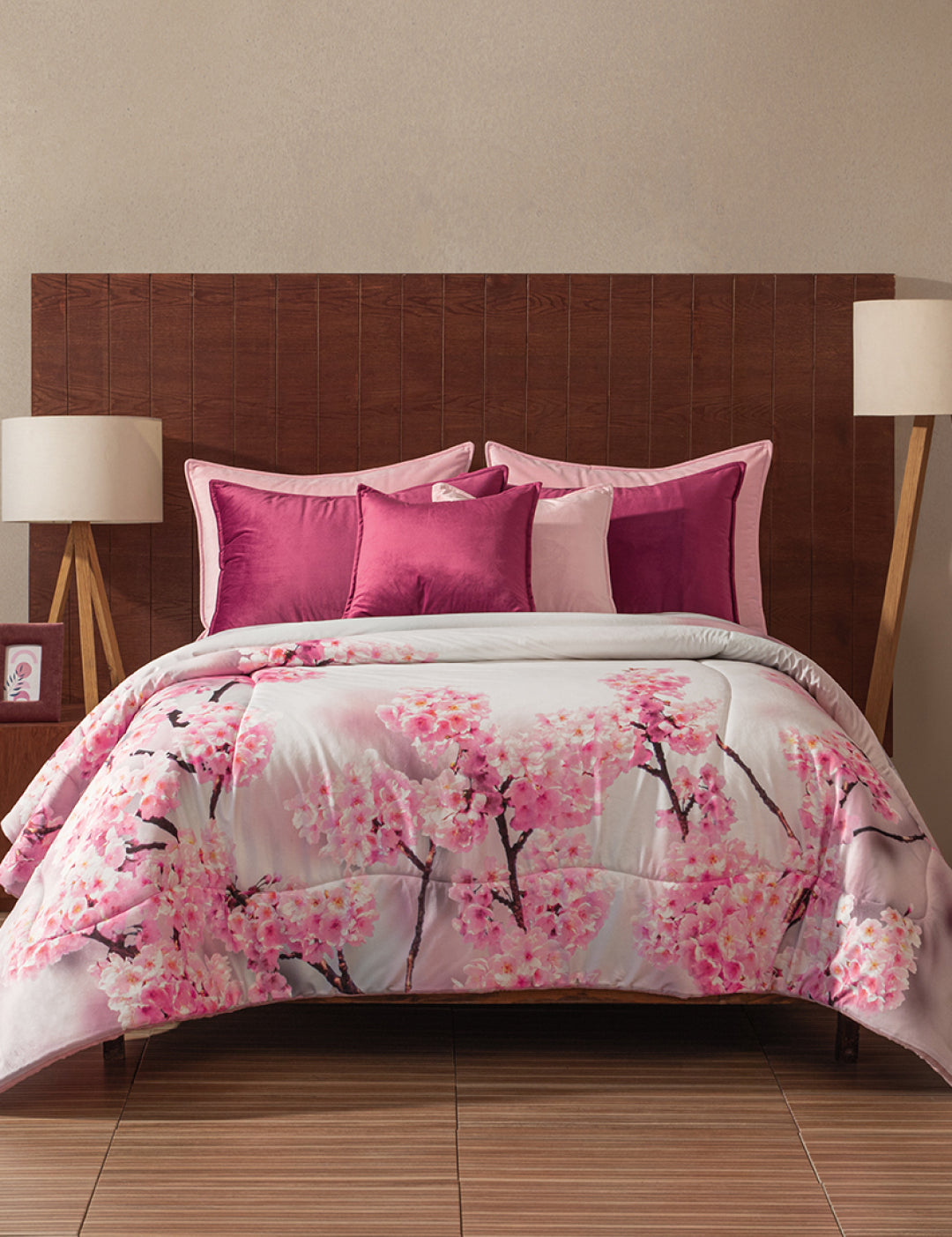 A bed showcases the Coordinado de Cobertor Digital Soft Florecer by Intima Hogar, adorned with pink cherry blossoms. Pink and magenta pillows accent the scene, complemented by a wooden headboard and modern bedside lamps.
