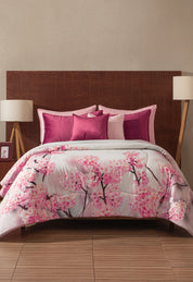 A bed showcases the Coordinado de Cobertor Digital Soft Florecer by Intima Hogar, adorned with pink cherry blossoms. Pink and magenta pillows accent the scene, complemented by a wooden headboard and modern bedside lamps.
