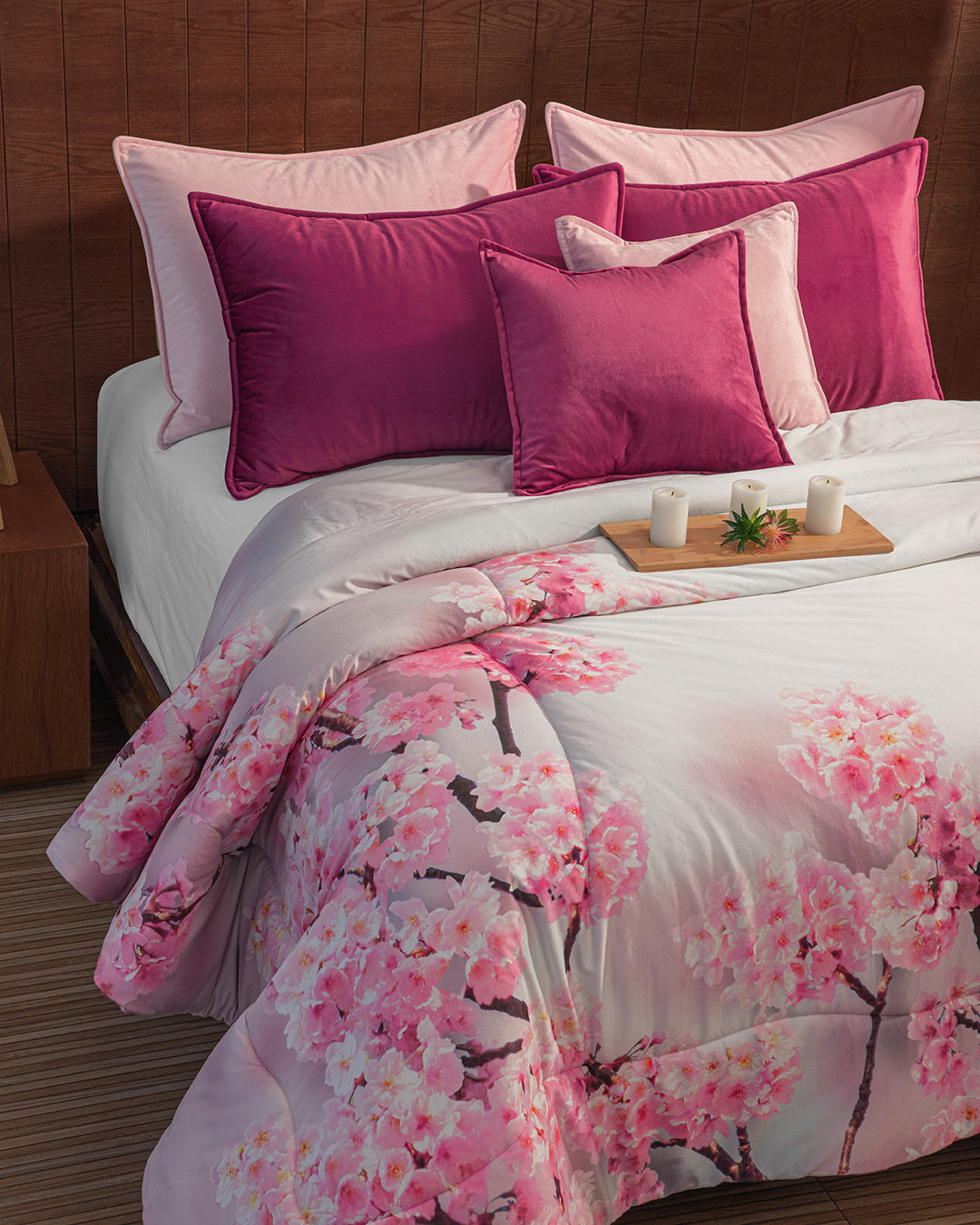A neatly made bed with pink and magenta pillows, the Coordinado de Cobertor Digital Soft Florecer by Intima Hogar featuring cherry blossoms, plus a wooden tray displaying three candles and a small green plant.