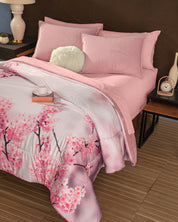 A neatly made bed features the Funda de Almohada Ensueño Rosa by Intima Hogar with matching pink pillows and a cherry blossom comforter. A round white cushion adds style, while a wooden nightstand holds a clock, lamp, and cup on a tray.