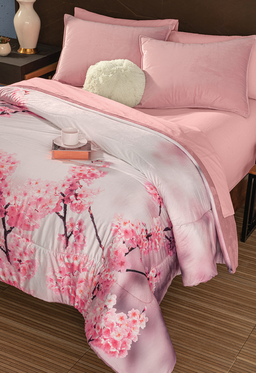 A neatly made bed with pink franela suave sheets, a round white pillow, and the Coordinado de Cobertor Digital Soft Florecer by Intima Hogar featuring cherry blossoms. A tray with a white cup and book sits on the bed, surrounded by warm-toned decor.