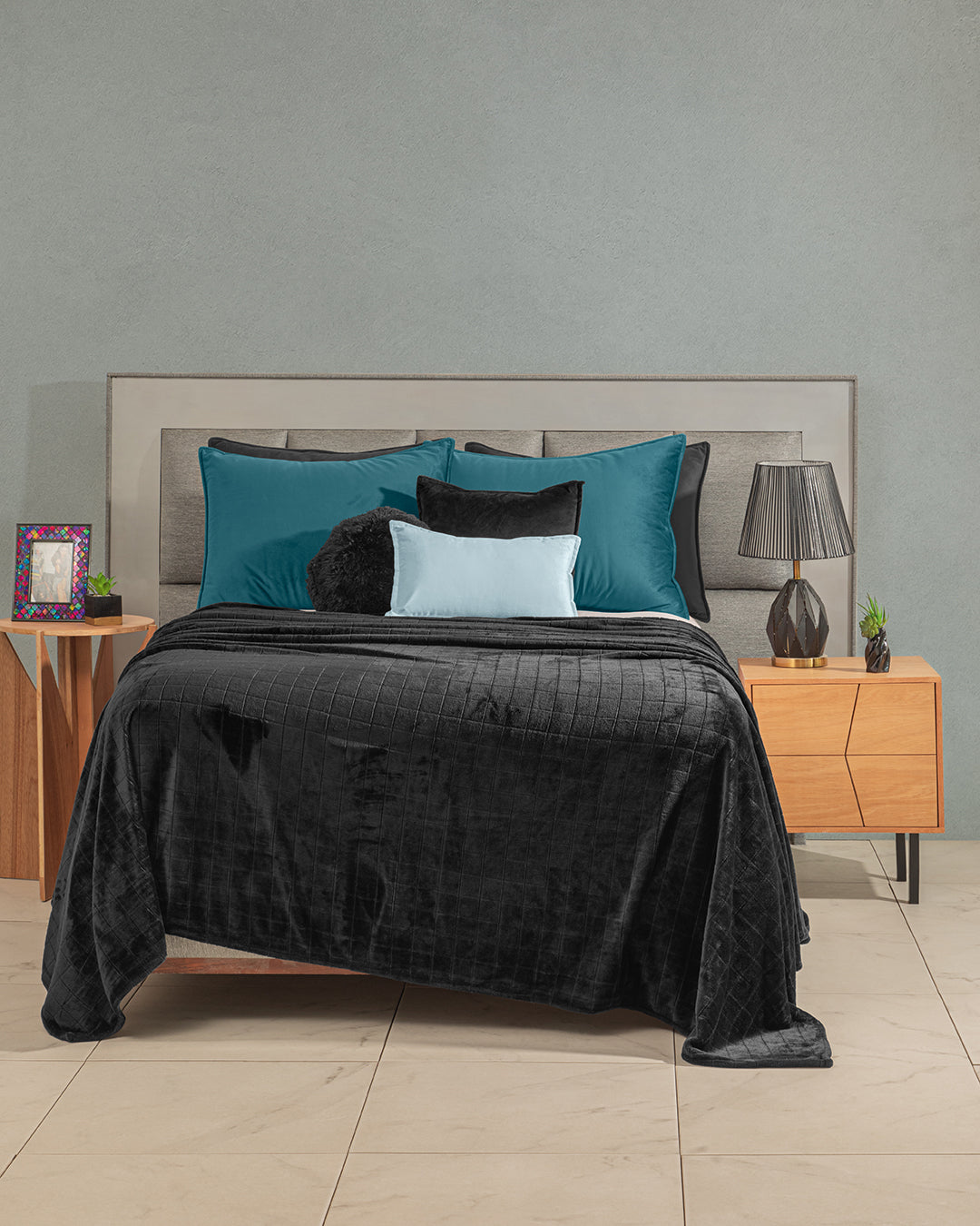 A modern dormitorio with a neatly made cama featuring Intima Hogar’s Funda de Almohada Ensueño Verde, teal and black pillows, a black quilted blanket, wooden nightstands with a lamp, framed picture, small plant, tiled floor, and light gray walls.