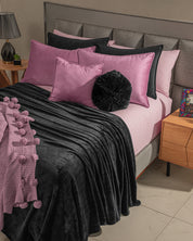 A modern cama de descanso with Intima Hogar's Funda de Almohada Ensueño Lila and a black textured blanket. The bed is next to a wooden nightstand with a lamp and colorful book, perfect for any habitación.
