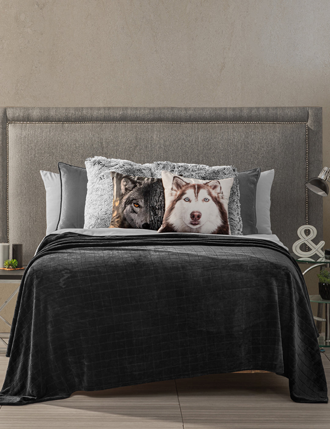 A modern bedroom features the Intima Hogar Coordinado de Cobertor Digital Soft Nebraska, a gray upholstered headboard, neat bedding, wolf and husky decorative pillows, plus side tables with lamps, small plants, and a decorative & sign.