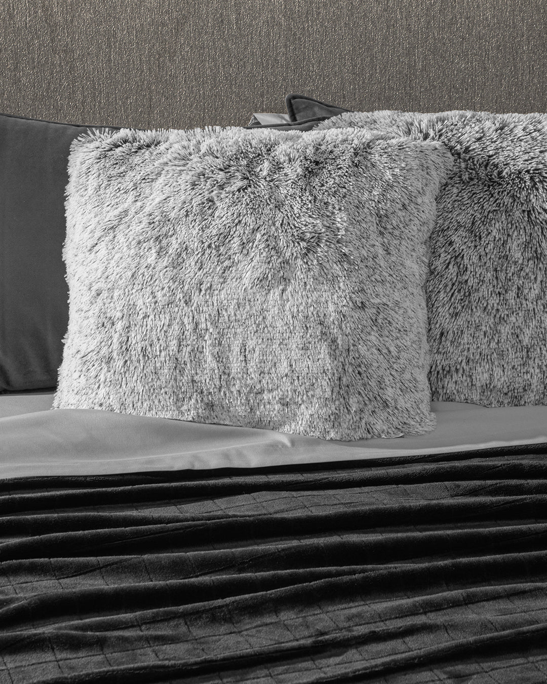 A close-up of a bed featuring the Juego de Cojines de Lujo Humo by Intima Hogar, paired with a gray sheet and dark textured blanket, set against a neutral wall.