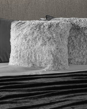 A close-up of a bed featuring the Juego de Cojines de Lujo Humo by Intima Hogar, paired with a gray sheet and dark textured blanket, set against a neutral wall.