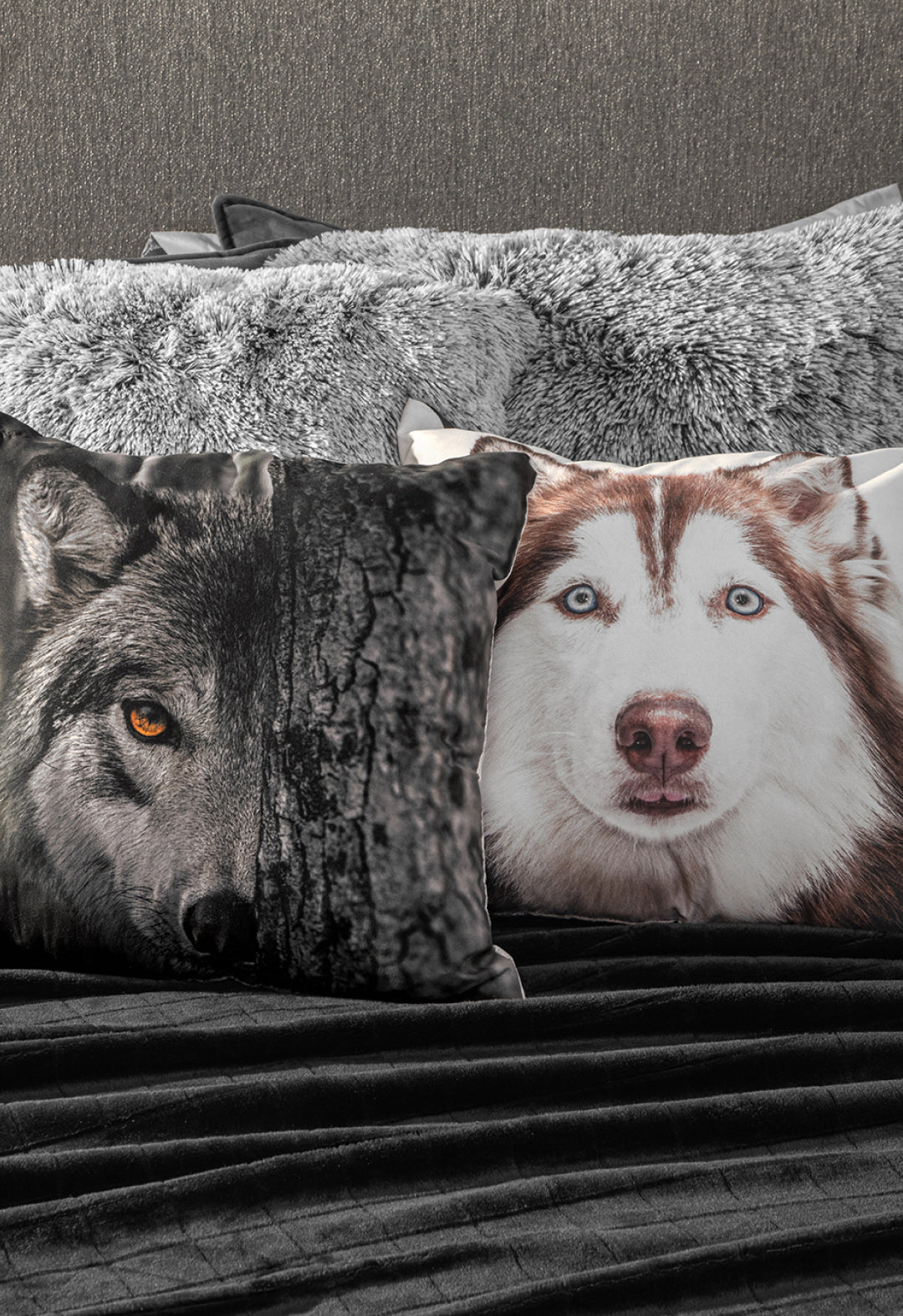 Two decorative pillows—one with a wolf’s face and one with a husky—rest on the Coordinado de Cobertor Digital Soft Nebraska by Intima Hogar, paired with gray faux fur pillows and a juego de sábanas Gris Oxford against a textured gray wall.