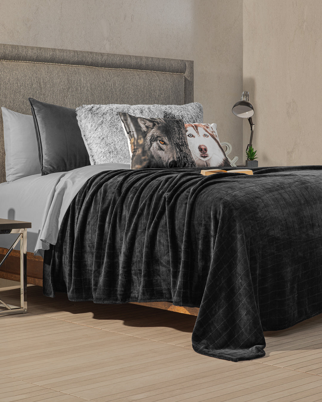 A modern bedroom features a bed with gray and white linens, a dark gray blanket, and decorative pillows—including the Cojín Estampado Lobo by Intima Hogar for visual interest. A reading lamp and a book sit on the bedside table.