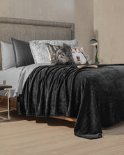 A modern bedroom features a bed with gray and white linens, a dark gray blanket, and decorative pillows—including the Cojín Estampado Lobo by Intima Hogar for visual interest. A reading lamp and a book sit on the bedside table.