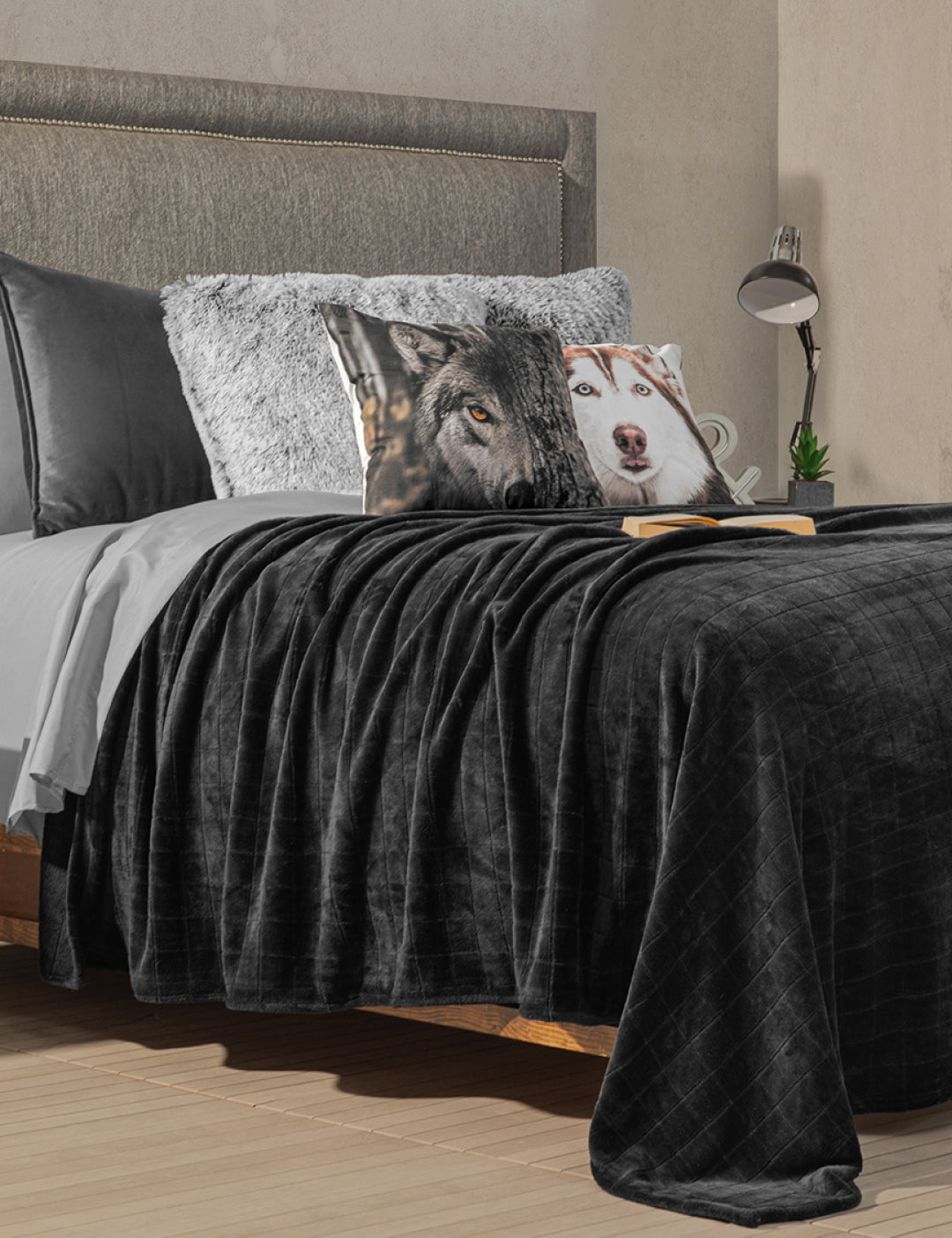 A neatly made bed with the Coordinado de Cobertor Digital Soft Nebraska by Intima Hogar and Juego de Sábanas Gris Oxford, plus a plush blanket, wolf and husky pillows. A book and eyeglasses rest on the bed; lamp and clock are on the nightstand.