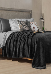 A neatly made bed with the Coordinado de Cobertor Digital Soft Nebraska by Intima Hogar and Juego de Sábanas Gris Oxford, plus a plush blanket, wolf and husky pillows. A book and eyeglasses rest on the bed; lamp and clock are on the nightstand.
