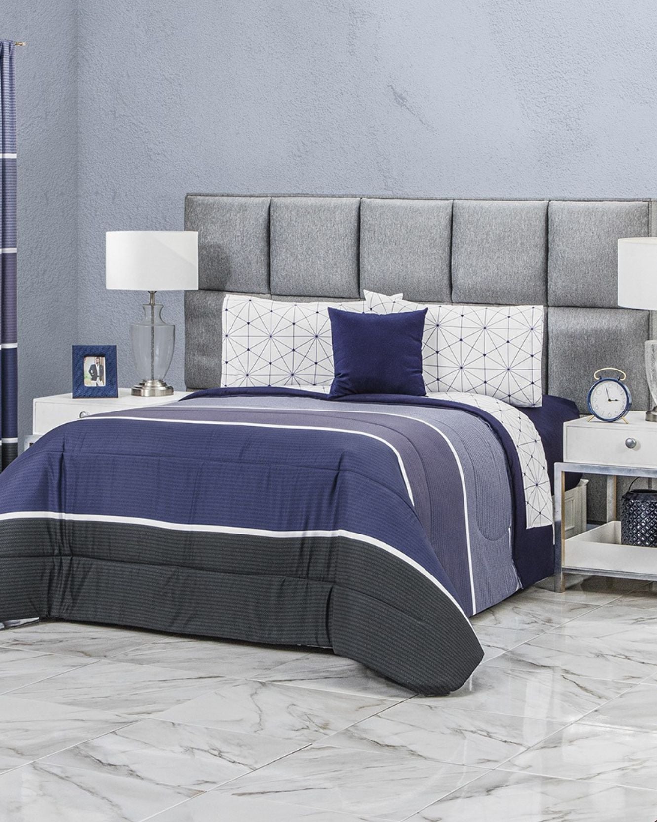 A modern bedroom with a gray upholstered headboard, Coordinado de Edredón Super Suave Aldo by Intima Hogar featuring blue and white geometric patterns, two white nightstands, lamps, a clock, and marble tile flooring.