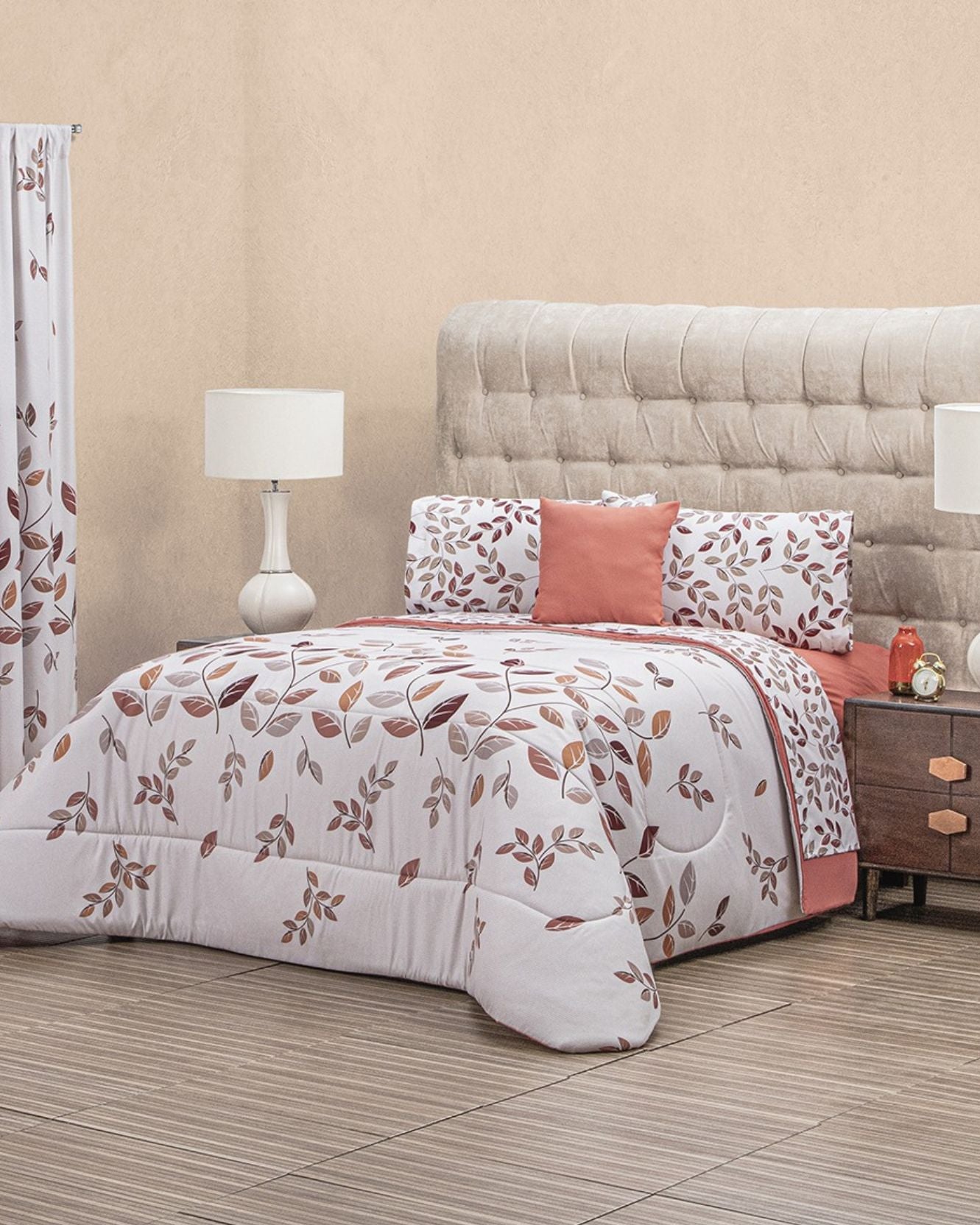 A neatly made bed features the Coordinado de Edredón Super Suave Antonela by Intima Hogar, with white bedding adorned with a brown and orange leaf design, matching pillowcases, coral accent pillows, bedside lamps, and a wooden nightstand in soft light.