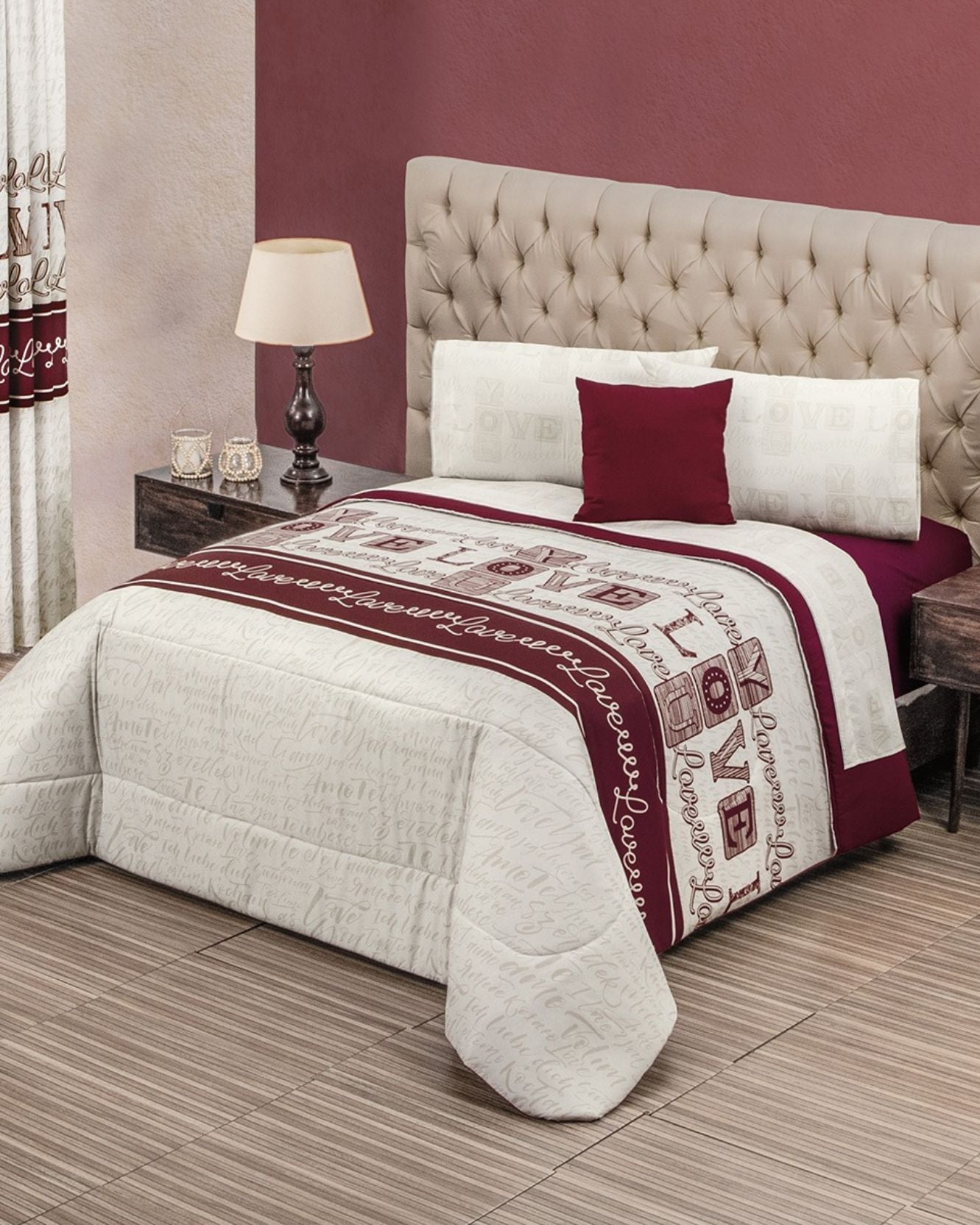 A neatly made bed with the Intima Hogar Coordinado de Edredón Super Suave Cupido, white and burgundy bedding with script and gift box patterns, three pillows, a beige tufted headboard, a lamp on a dark nightstand, and red and beige walls.