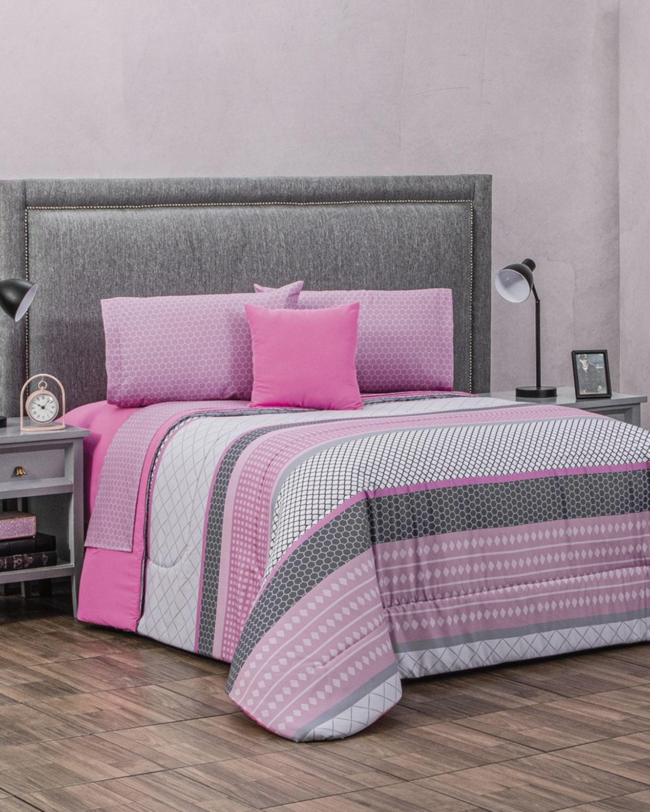 A neatly made bed with a gray upholstered headboard features the Coordinado de Edredón Super Suave Daria by Intima Hogar, including a pink and gray patterned edredón, matching cojín, and game de sábanas. Two bedside tables with lamps stand on wooden flooring.