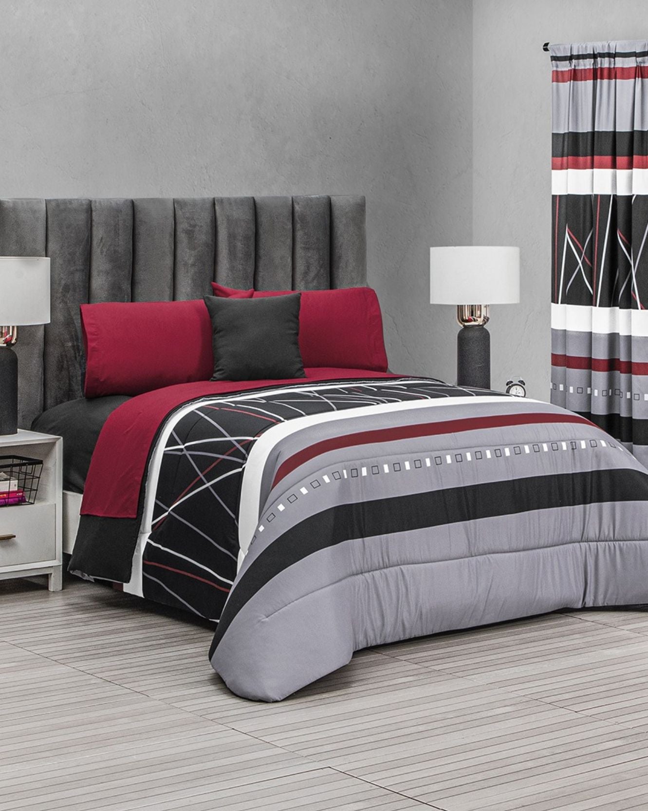 A modern bedroom featuring Intima Hogar’s Coordinado de Edredón Super Suave Gael, with red, black, white, and gray geometric bedding, matching pillows, a gray upholstered headboard, bedside tables with lamps, and striped curtains.