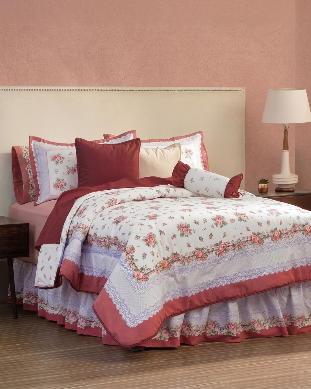 A neatly made bed showcases the Coordinado de Edredón Galicia by Intima Hogar in pink and white, with matching pillows, beige headboard, nightstand with lamp and plant. The room has a light pink wall and wood flooring.