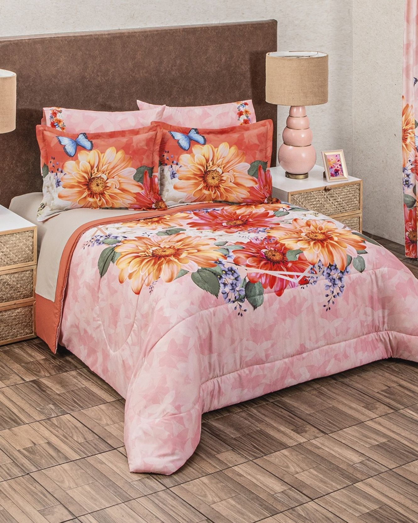 A bed is styled with the Coordinado de Edredón Super Suave Mandarina by Intima Hogar, showcasing a pink floral duvet with orange flowers, green leaves, and blue butterflies. Two matching decorative shams rest against a brown headboard.