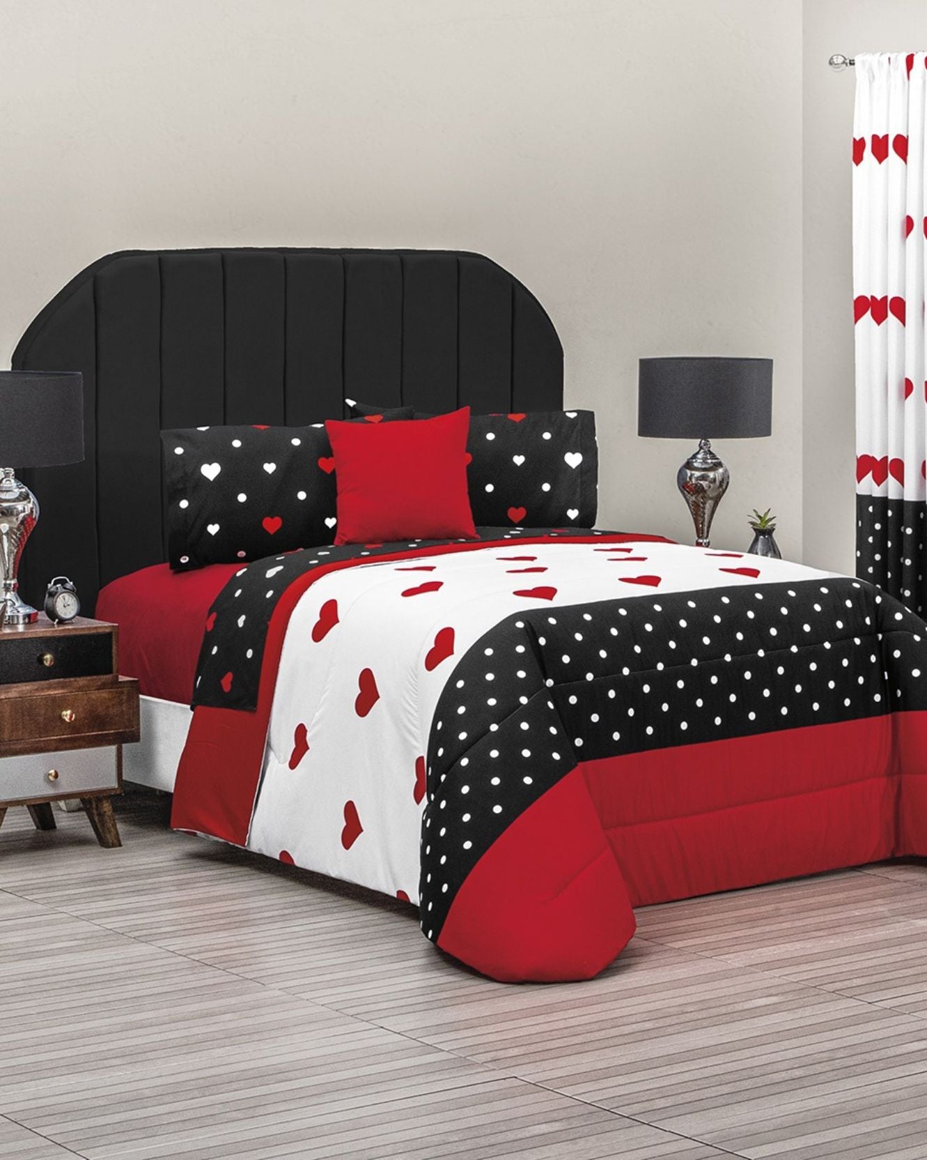 A neatly made bed showcases the Coordinado de Edredón Super Suave Nina by Intima Hogar, featuring black, white, and red heart and polka dot patterns. The room includes a black headboard, lamps, a bedside table, and a red heart curtain.