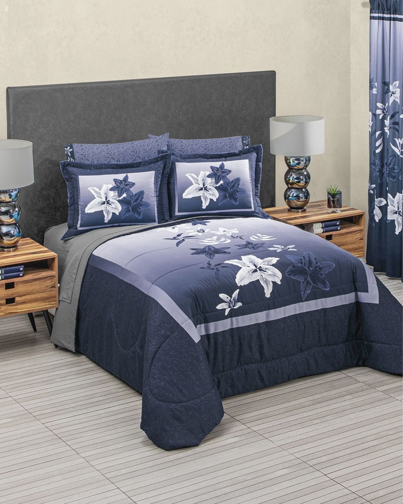 A neatly made bed showcases the Intima Hogar Coordinado de Edredón Super Suave Rita, complete with matching pillow shams, fundas decorativas, and decorative pillows. Wooden nightstands with lamps flank the bed, and a blue floral curtain accents the window.