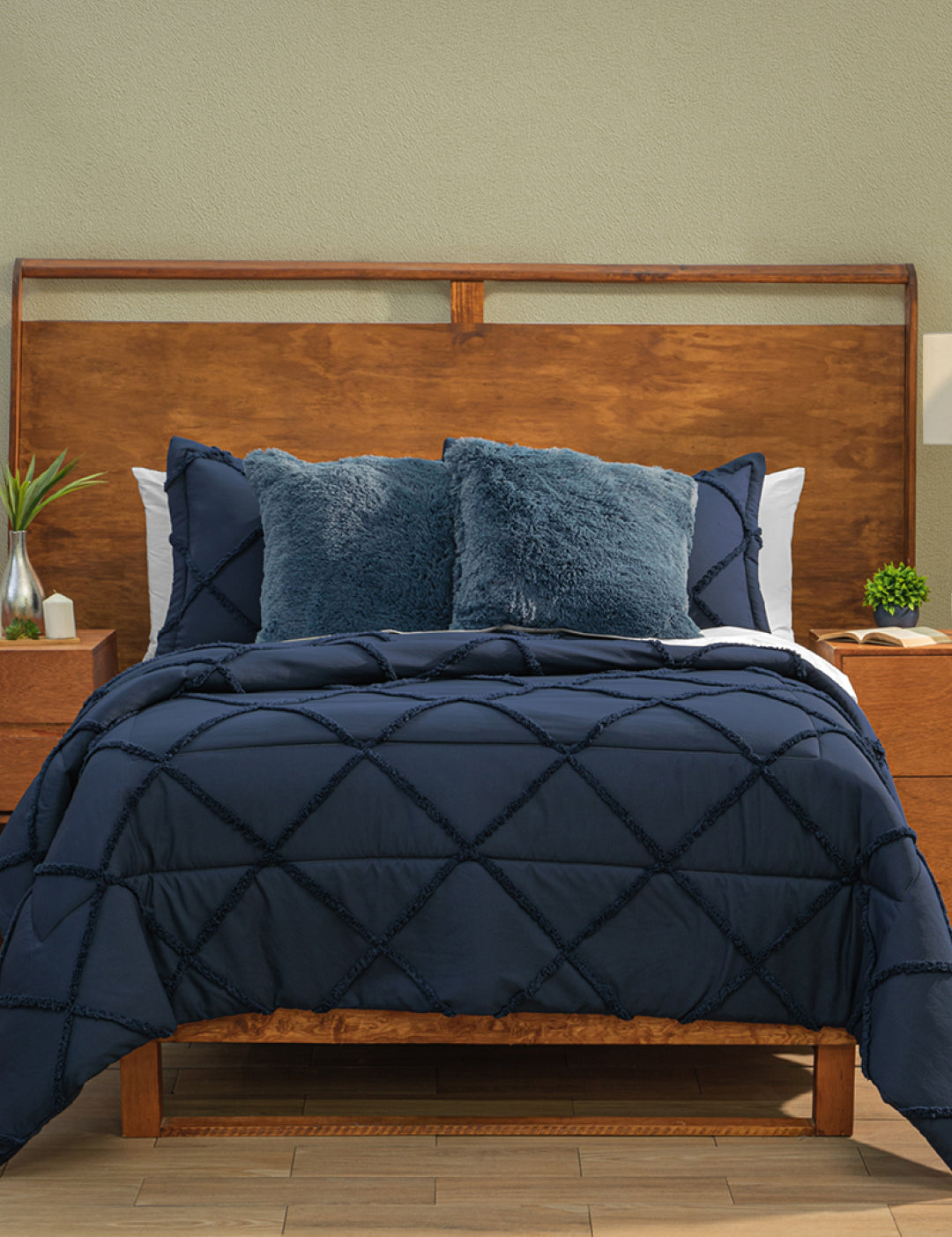 A neatly made bed features the Intima Hogar Coordinado de Edredón Luxury Seúl in dark blue with a diamond pattern, matching pillows, two textured navy accent pillows, a wooden headboard, light green wall, and nightstands with plants and lamps.
