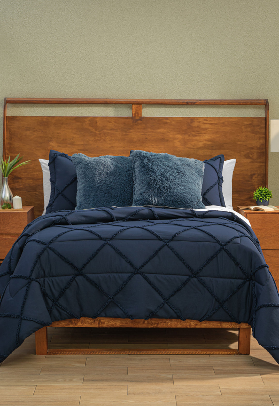 A neatly made bed features the Intima Hogar Coordinado de Edredón Luxury Seúl in dark blue with a diamond pattern, matching pillows, two textured navy accent pillows, a wooden headboard, light green wall, and nightstands with plants and lamps.