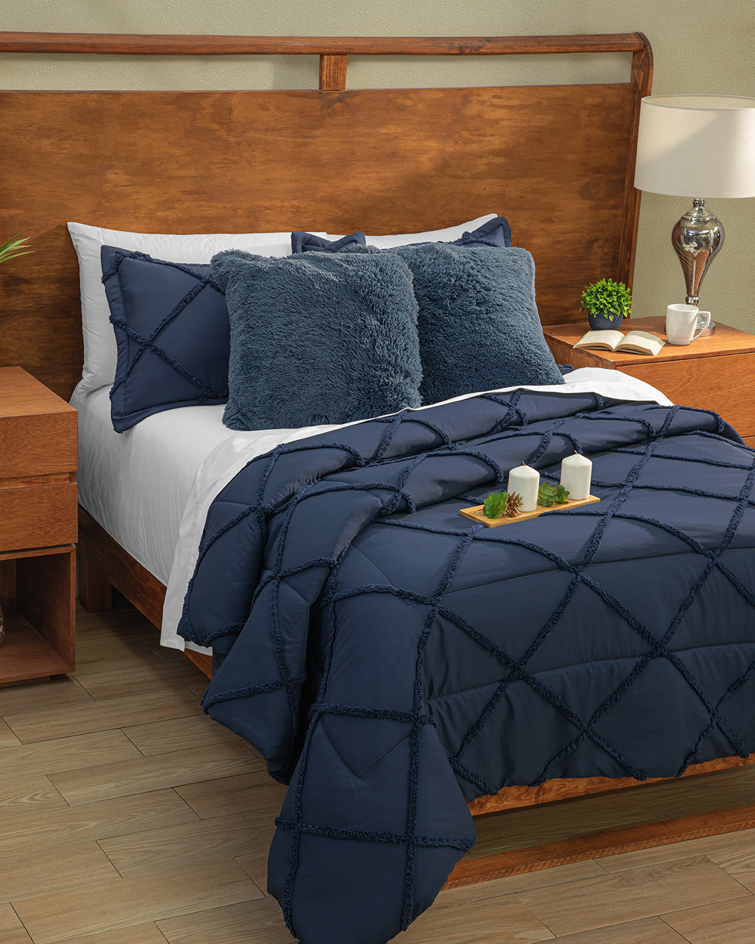 A neatly made bed features a navy blue textured comforter, Intima Hogar Cojines de Lujo Bruselas, white sheets, and a wooden headboard. A tray with candles and greenery sits on the bed, with wooden nightstands and a lamp beside it.